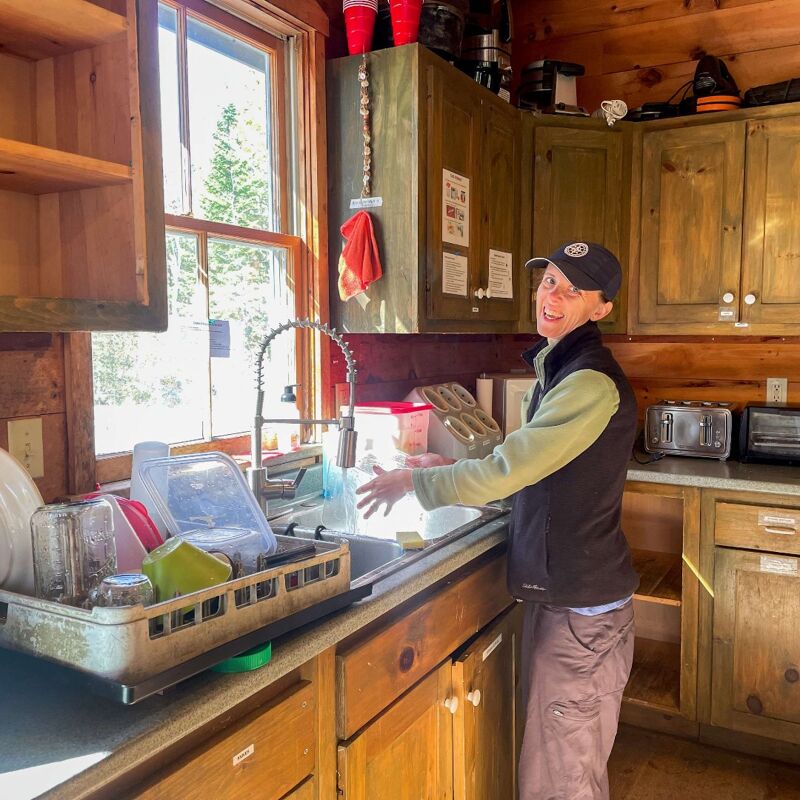 The image shows a person in a rustic kitchen. They are standing at a sink, possibly washing dishes. The kitchen has wooden cabinets and a window that lets in natural light. There are various items on the counter, including a dish rack and containers. The person is smiling, suggesting a positive mood.